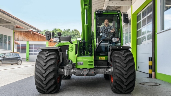 Widok Fendt Cargo T955 od przodu z wysuniętym ramieniem teleskopowym i kierowcą w kabinie na placu roboczym.