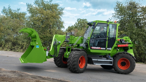 Rolnik stoi na podwórzu obok Fendt Cargo T955 z otwartym chwytakiem do kiszonki; w tle widać drzewa.