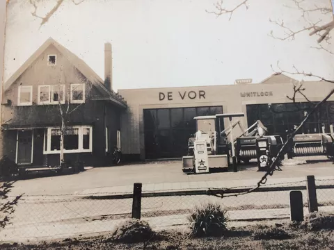 Historical black and white photo of a house with an attached workshop and agricultural machinery in the foreground, de Vor's first dealership in the 1950s