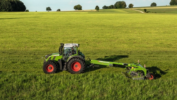 Fendt Slicer trailed with transport chassis on a large meadow while mowing. Scattered trees and bushes can be seen in the background.