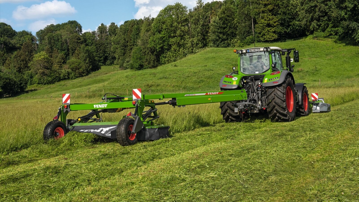 A farmer driving a tractor in a meadow and mowing with a towed Fendt Slicer mower.