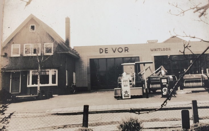 Historical black and white photo of a house with an attached workshop and agricultural machinery in the foreground, de Vor's first dealership in the 1950s